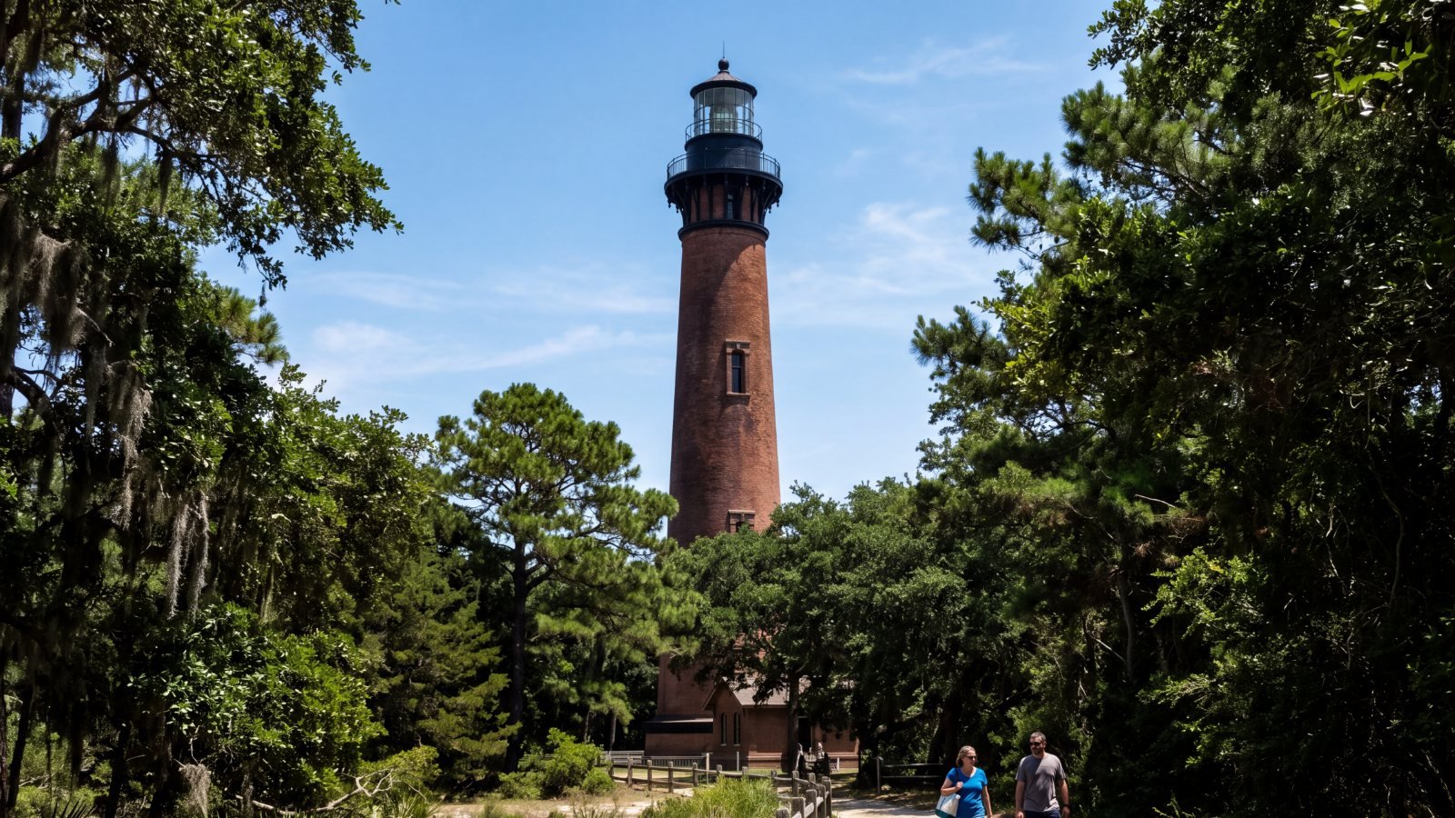 A bare red-brick lighthouse rising above live oaks over a shaded lane, with two people walking toward it
