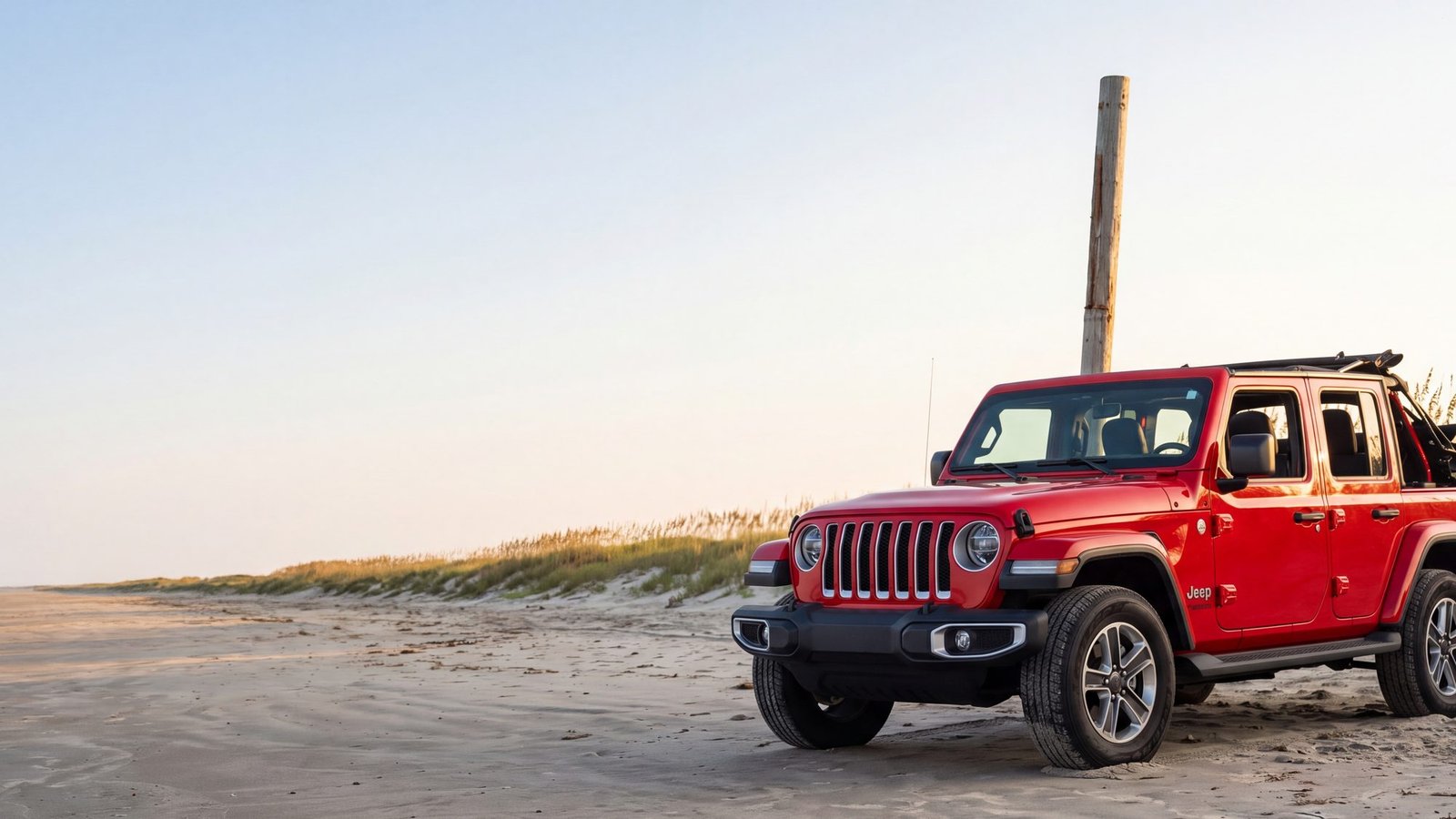 A red four-door Jeep Wrangler with its soft top folded back, parked on hard-packed sand at the top of an empty Outer Banks beach beside a weathered dune-crossing post