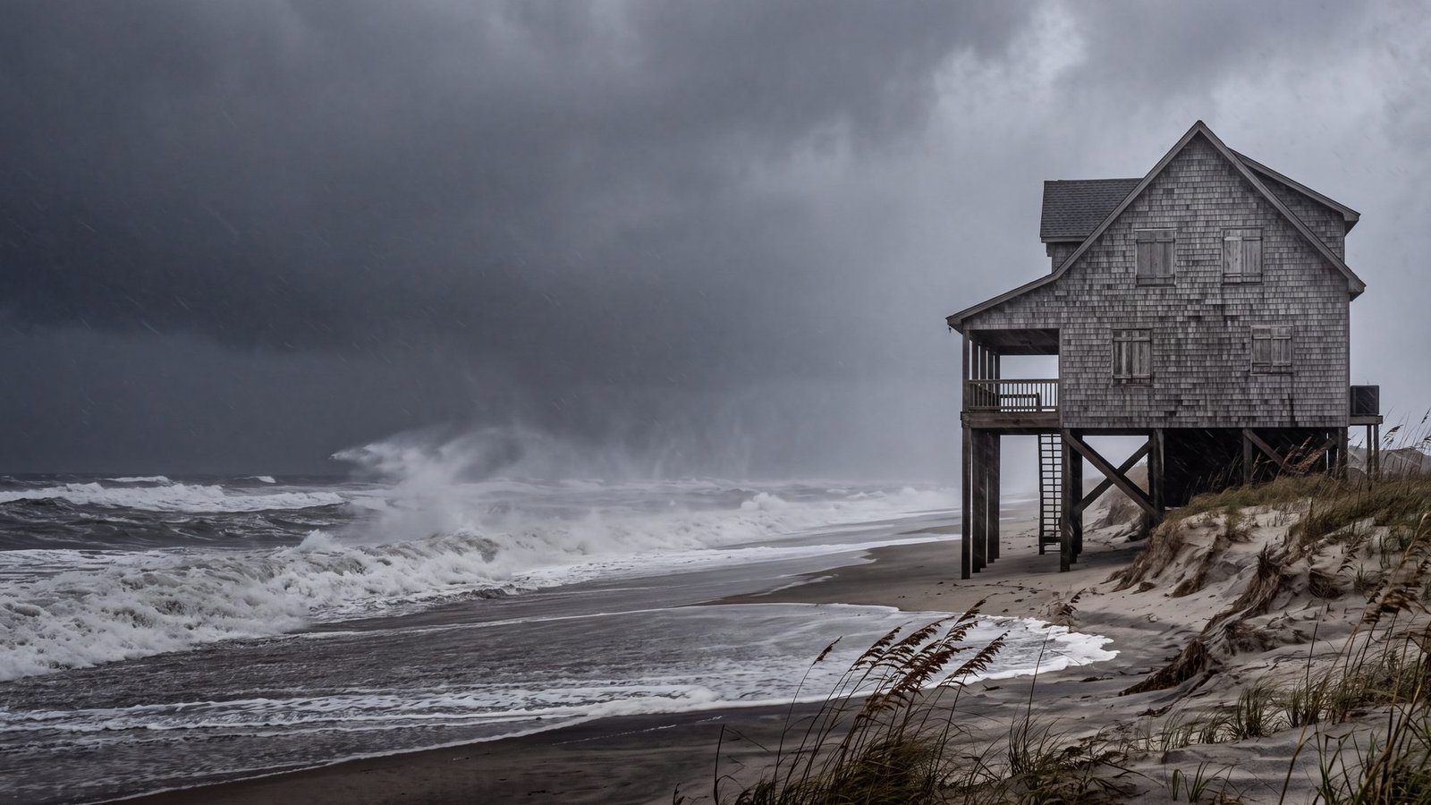 Storm surf and blown spray in front of a shuttered cedar-shingle cottage on pilings under a dark sky